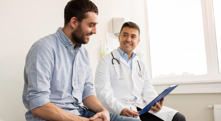 Young man patient sat with a male doctor on a bed in a white room. Both smiling supportively.